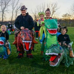 A family stands on a grassy field dressed in dinosaur costumes. Two adults wear inflatable T. rex outfits, while three children wear smaller dinosaur suits. The sunset and sparse trees are visible in the background.
