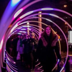 People walk under a colorful, illuminated tunnel with glowing rings in pink and purple. A woman in a coat is in the foreground, followed by others taking photos. The scene is lively and vibrant, highlighting an urban light installation at night.