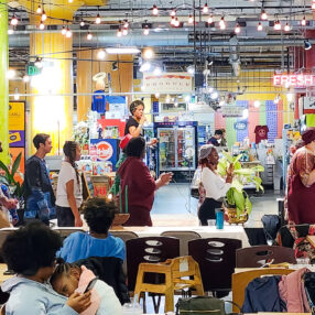 A lively scene at the Kwanzaa Celebration at Midtown Global Market with people dancing in a group. A singer performs on a small stage amid colorful decor and string lights. In the foreground, a person cradles a resting child. The atmosphere is vibrant and festive.