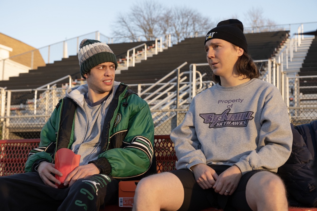 Pete Davidson and Paul Dano in Dumb Money sitting on bleachers.