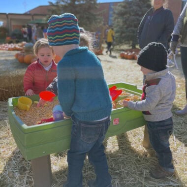 At the Kids at Pumpkin Patch at Our Lady of Peace Minneapolis, three children in warm clothing play at a green sensory table filled with corn kernels outdoors. There are pumpkins in the background and a few adults nearby.