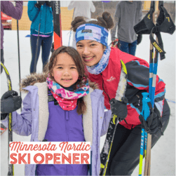 Two smiling people pose outdoors with ski gear during the Minnesota Nordic Ski Opener event. The older person wears a red jacket and blue headband, while the younger one is in a purple coat and colorful scarf. Snow-covered ground is visible.