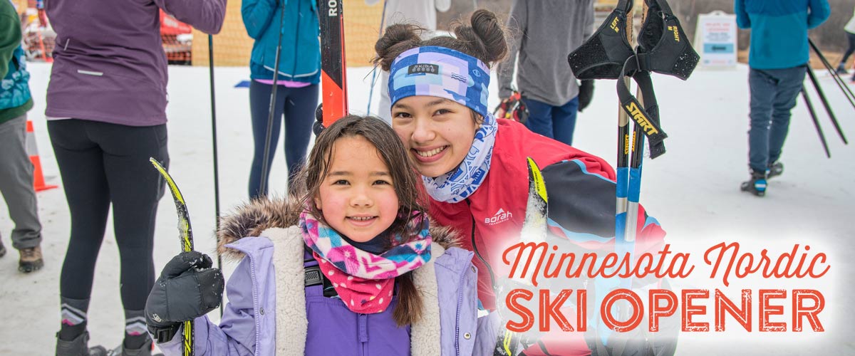 A smiling adult and child in colorful winter clothing and ski gear pose outdoors at the Minnesota Nordic Ski Opener. Other participants can be seen in the background, with skis and poles visible.