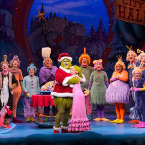 The image shows a colorful stage performance with characters dressed as whimsical, festive creatures. A green character in a Santa outfit is at the center, surrounded by a joyful group in bright costumes, under a "City Hall" sign.