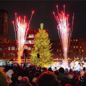 A large, brightly lit Christmas tree stands in a busy square at night, surrounded by a crowd. Fireworks light up the sky on either side of the tree, creating a festive and celebratory atmosphere.