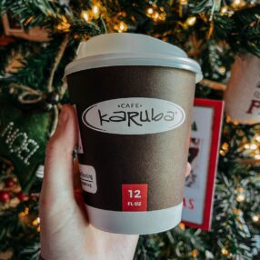 A hand holds a 12 fl oz Karuba coffee cup from KwikTrip in front of a decorated Christmas tree with lights.