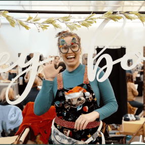 A person wearing colorful Christmas glasses, a teal shirt, and a festive apron is smiling and waving behind a booth with large cursive text at Minnesota Merry Market. The setting appears lively, with various people in the background engaged in activities.