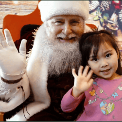A man dressed as Santa Claus and a young girl are sitting together, both smiling and waving at the camera at Minnesota Merry Market. The girl is wearing a pink shirt with colorful designs. The background is festive and resembles a wooden texture with Christmas decorations.