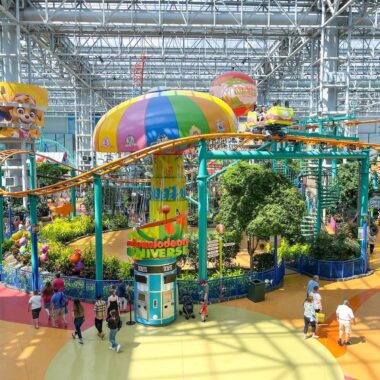 Colorful indoor amusement park with a roller coaster winding around a large, playful central structure adorned with cartoon-themed decorations. Visitors are walking around and enjoying the lively atmosphere and attractions.