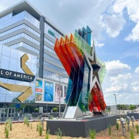 A large, colorful star sculpture stands outside the Mall of America. The mall's glass facade reflects the sky, and banners advertising an aquarium are visible. The scene is set against a partly cloudy blue sky.