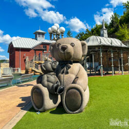 A large stone sculpture of two teddy bears, one sitting with a bow tie, the other playing a saxophone, is set on a green lawn in Teddy Bear Park in Stillwater Minnesota. Behind them, there are a red building, a gazebo, and a clock tower. The sky is clear with scattered clouds.