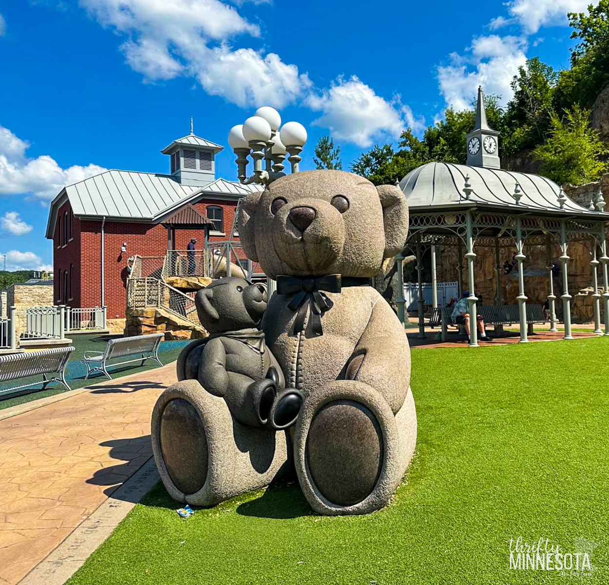 A large stone sculpture of two teddy bears set on a green lawn in Teddy Bear Park in Stillwater Minnesota. Behind them, there are a red building, a gazebo, and a clock tower. The sky is clear with scattered clouds.