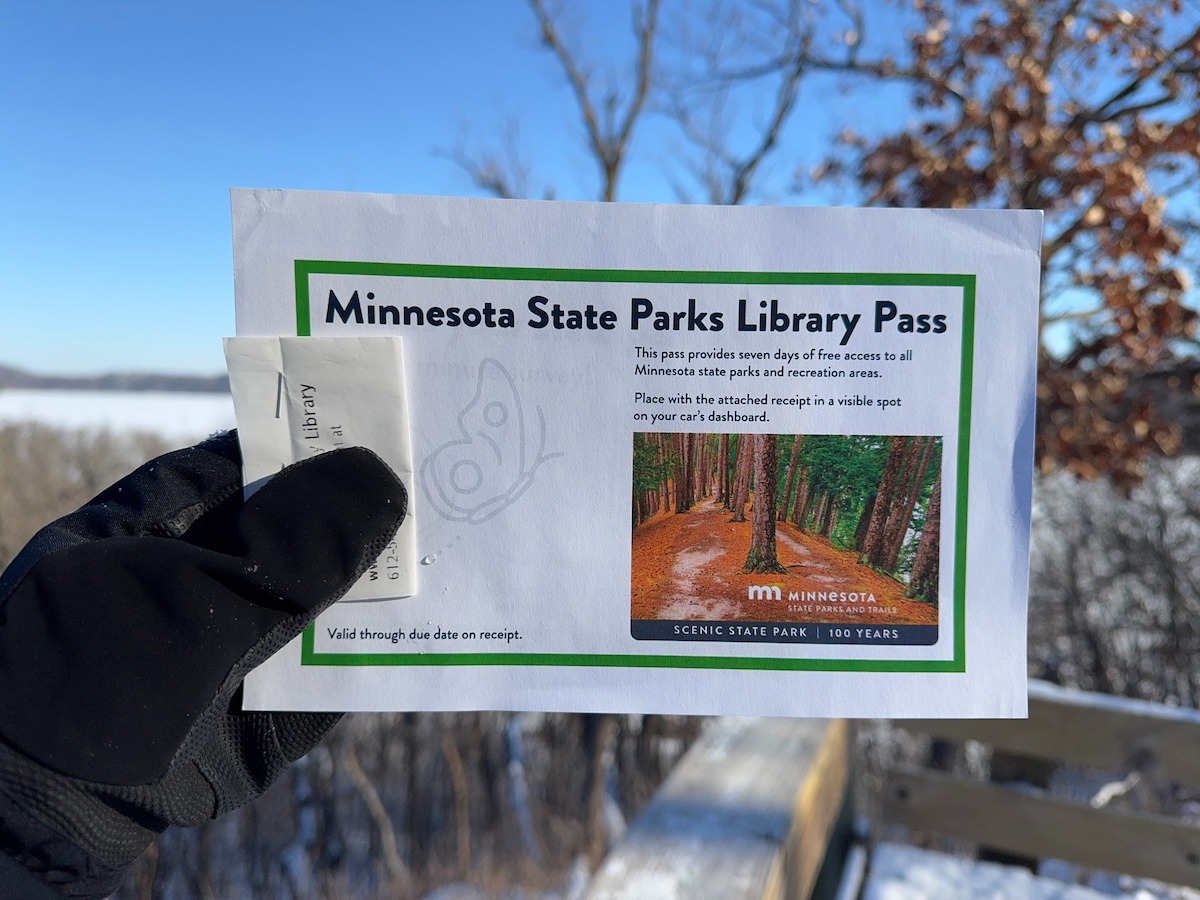A gloved hand holds up the MN State Parks Library pass in the foreground with a snowy and blue-skied day in the background.