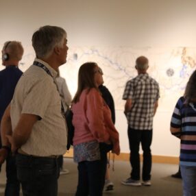 People standing in an art gallery, observing a large abstract artwork on the wall. Some are wearing headphones. The room is softly lit, creating a contemplative atmosphere.