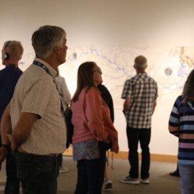 People standing in an art gallery, observing a large abstract artwork on the wall. Some are wearing headphones. The room is softly lit, creating a contemplative atmosphere.