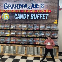 A child stands smiling in front of a large candy display labeled "Grandpa Joe's World Famous Candy Buffet." The display features a variety of colorful candies in containers. Two rubber ducks decorate the top. The child holds a box of candy.