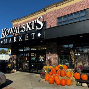 A storefront of Kowalski's Market with a brick facade and large windows. The entrance is decorated with an assortment of pumpkins, squash, and fall flowers. Shadows from trees are visible on the ground.
