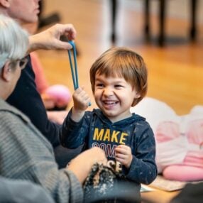 A smiling child in a navy hoodie plays with a blue rubber band, surrounded by adults. One adult, with short white hair, sits nearby. The room has wooden flooring and baskets visible on the floor.