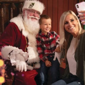 A woman takes a selfie with a young boy sitting on Santa's lap. Santa is dressed in his traditional red suit, and everyone is smiling. Festive lights decorate the foreground, adding a cheerful holiday atmosphere.