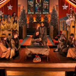 A Christmas nativity play with children in costumes as shepherds, angels, and other biblical figures. A manger is at the center. The backdrop includes stained glass, stars, and Christmas trees adorned with lights.