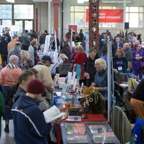 A bustling book fair with attendees browsing various stalls filled with books. Authors are signing books at a dedicated section, and people are engaged in conversations. The atmosphere is lively and vibrant with a diverse crowd.