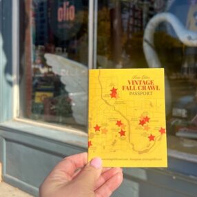 A hand holds a "Vintage Fall Crawl Passport" brochure with a map and red stars, in front of a shop window.