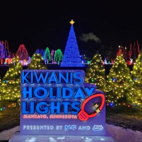 A nighttime holiday display featuring a lit sign reading "Kiwanis Holiday Lights, Mankato, Minnesota" in front of vibrant Christmas lights. A tall, illuminated Christmas tree and colorful light-wrapped trees are visible in the background.