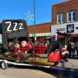 A decorated float in the Aitkin Fish House parade features Santa and others in red plaid outfits sitting in a makeshift sleigh made from a fishing boat and an ice shack with a black cover labeled "ZZZ." The sleigh is adorned with festive decorations and a sign reading "What Moose?.