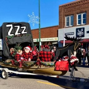 A decorated float in the Aitkin Fish House parade features Santa and others in red plaid outfits sitting in a makeshift sleigh made from a fishing boat and an ice shack with a black cover labeled "ZZZ." The sleigh is adorned with festive decorations and a sign reading "What Moose?.