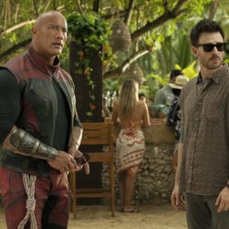 Two men stand in an outdoor setting with trees and a bar in the background. One wears a red and black costume, and the other is in casual attire with sunglasses. People are gathered around the bar.