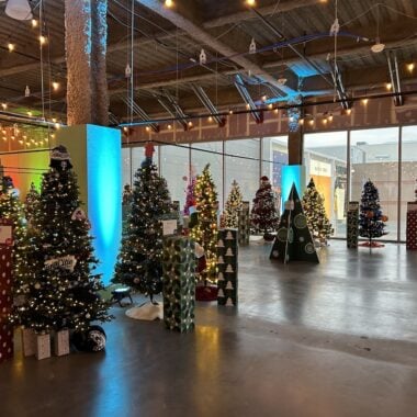 A festive indoor scene with a variety of decorated Christmas trees arranged in rows. Some trees are lit, and there are wrapped gift boxes at their bases. The room is illuminated with hanging string lights and ambient lighting.