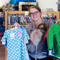 A person smiles while holding a baby in a carrier at their chest. They are holding up a blue polka-dotted onesie and a green jacket. The background shows shelves with books and other colorful clothing.