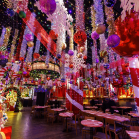 Jingle Bar Minneapolis decorated extensively with holiday decor, including colorful lights, large ornaments, and tinsel. Red and white striped columns resemble candy canes, creating a festive and lively atmosphere. Tables are set for dining.