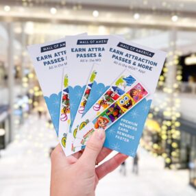 A hand holds several "Mall of America" pamphlets featuring "Earn Attraction Passes & More" in a brightly lit indoor shopping environment with blurred store backgrounds and decorative lights.
