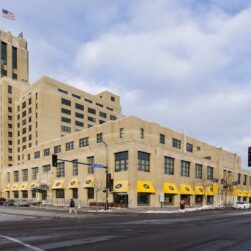 A tall, historic building with a beige facade and clock tower stands at a street intersection. Yellow awnings line the ground floor shops. An American flag flies atop the tower. Snow is on the ground and a few pedestrians are walking by.