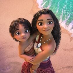 An animated scene of two characters with curly hair standing on a beach. The older character, wearing a necklace, is holding a younger one, near the shore where the water meets the sand. Both are looking upward with smiles.