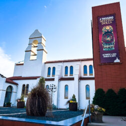A white historical building with red-tiled roofs under a blue sky. A banner on the tower advertises "A Thousand Nestling Dolls" exhibition from October 5, 2024, to March 9, 2025. Potted plants and a bare tree are in the foreground.