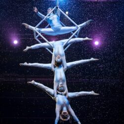 A group of acrobats in white costumes perform an aerial act, stacked in a vertical formation. They are suspended under a lit stage with blue and purple spotlights, surrounded by falling snow-like particles.