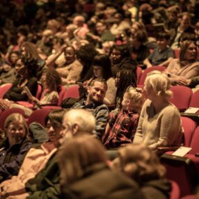 A full auditorium with people of different ages seated in red chairs, many engaged in conversations or looking at the stage. The atmosphere appears lively and anticipatory.