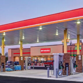 Holiday Stationstore gas station with a large red canopy, multiple fueling stations, and a convenience store in the background. The store sign reads "Holiday." The area is clean and well-lit, and there are some products stacked outside the store.