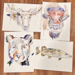 Four watercolor paintings on a wooden floor depict animals: a moose, a buffalo, a bear, and a fish. Each painting uses soft, blended colors to create a fluid, artistic representation of the animals.