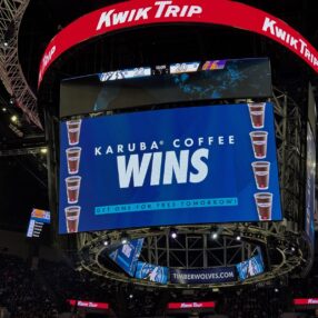 Target Center scoreboard displays "Karuba Coffee Wins" with an offer for free coffee from the KwikTrip Kwik-Tac-Toe Game. It shows a series of coffee cup images. The scoreboard is surrounded by advertisements and there's a crowd watching the Timberwolves game below.