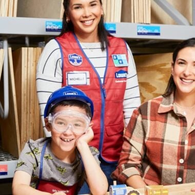 Three people in a workshop: a smiling woman in a plaid shirt sitting next to a boy wearing a blue helmet and safety goggles, and a woman in a red apron standing behind them. They are surrounded by wooden panels and tools.