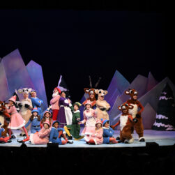 A colorful stage performance featuring actors in reindeer, elf, and snowman costumes against a snowy mountain backdrop. The scene is lively, with performers posing and smiling under bright stage lights.