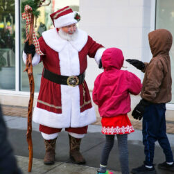 A person dressed as Santa Claus stands outdoors, holding a wooden staff decorated with greenery and ribbon. Two children in hooded jackets face Santa, seemingly engaged in conversation or greeting, on a paved walkway in front of a building.