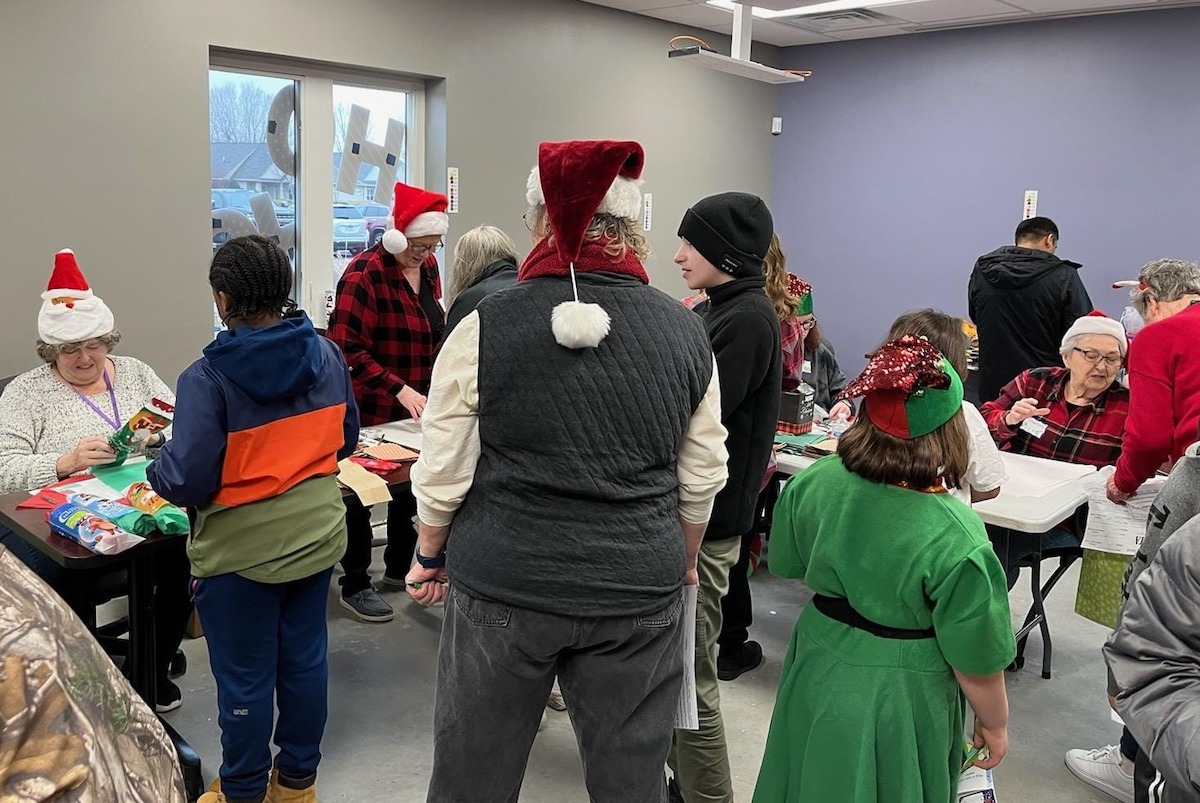 A group of people, some wearing festive hats, gather in a room with tables for Secret Holiday Shop. The setting is lively and cheerful.
