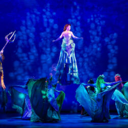 A theatrical performance of The Little Mermaid at the Ordway theater with actors in vibrant costumes on stage. Ariel is at center in a flowing, blue-green dress appears swiming, surrounded by performers in aquatic-themed outfits. The lighting creates an underwater effect with shades of blue and green.