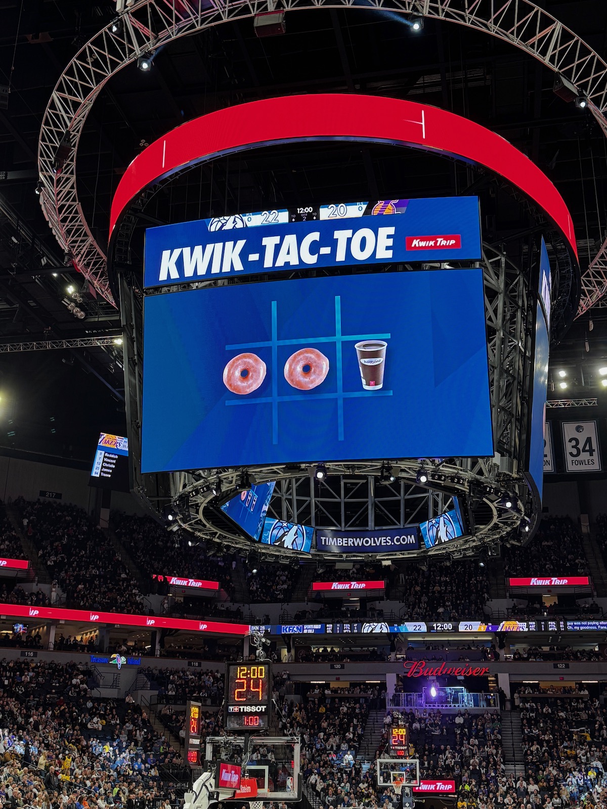 Target Center Minneapolis with a large screen displaying the KwikTrip Tic-Tac-Toe game featuring donuts and Karuba coffee as symbols. The audience watches, and the scoreboard shows Timberwolves branding and a score of 24-12.