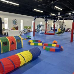 A spacious indoor play area with colorful play equipment on blue mats, including a tunnel, foam pits, swings, and climbing structures. The room has a section for arts and crafts with tables and chairs. Bright lighting and a vibrant atmosphere.