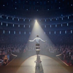 A performer stands on stage under a spotlight, arms outstretched, facing a large audience seated at tables in a grand theater. The crowd is watching attentively, filling the space with an atmosphere of anticipation and excitement.